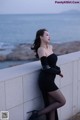 A woman in a black dress leaning against a wall by the ocean.